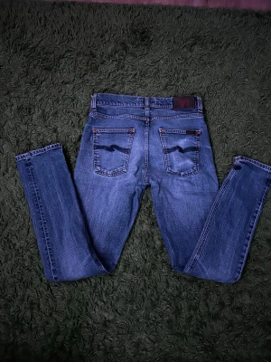 Blå jeans från Nudie Jeans Co - Säljer ett par klassiska blå jeans från Nudie Jeans Co med raka ben och snygga kontrastsömmar i orange. Bakfickorna har den ikoniska vågiga sömmen och jeansen är gjorda i slitstark denim. Perfekta för dig som gillar stilrena och tidlösa jeans. W30/L32