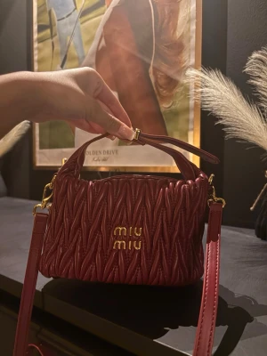 Vinröd quiltad handväska från Miu Miu - Supersnygg vinröd handväska från Miu Miu i quiltat läder med guldiga detaljer och logga framtill. Väskan har både axelrem och handtag, perfekt för att bära på olika sätt. Den lyxiga looken och den djupa färgen gör den till ett statement-piece. Tror den är äkta men Kan ej garantera autencitet. Dustbag medföljer!