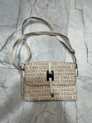 Beige axelväska med textmönster - Trendig beige axelväska med justerbar rem och textmönster där det står 'CHANGE TIME COLORS'. Väskan har ett spänne framtill och flera fack med dragkedja inuti, perfekt för att hålla ordning på dina grejer. Materialet ser ut att vara konstläder.