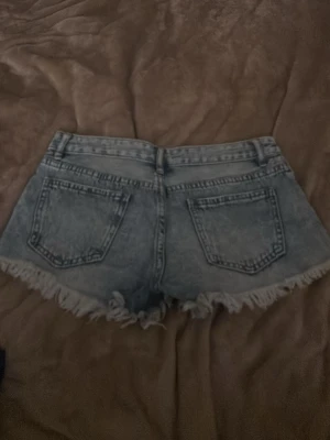Blå jeansshorts med fransig kant - Säljer ett par ljusblå jeansshorts med fransig, rå kant och klassisk femficksmodell. Shortsen har slitna detaljer och hög midja, perfekta för en avslappnad och trendig look. Inget synligt märke på shortsen.