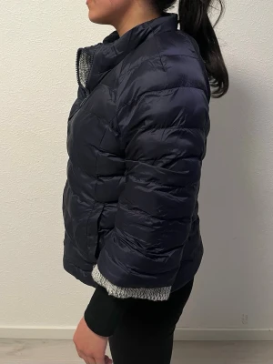 Trekvartsärmad jacka - Säljer en mörkblå pufferjacka med dragkedja och hög krage. Jackan har en unik design med en grå stickad insida som syns vid ärmslut och krage. Perfekt för kalla vinterdagar och ger både värme och stil. Materialet är quiltat syntet och stickad ullblandning.