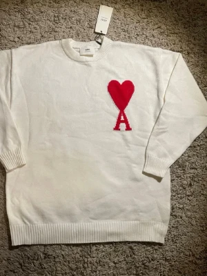 Vit stickad tröja från Ami Paris - Säljer en vit stickad tröja från Ami Paris med det ikoniska röda hjärtat och bokstaven A på bröstet. Tröjan har rund halsringning, ribbade muddar och är tillverkad i mjukt material som känns lyxigt och trendigt.