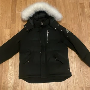 Svart Moose Knuckles med pälskantad huva - Snygg svart dunjacka med stor huva och vit pälskant. Jackan har flera fickor med dragkedja och tryckknappar samt en patch på ärmen. Perfekt för kalla vinterdagar och ger en riktigt cool streetstil.