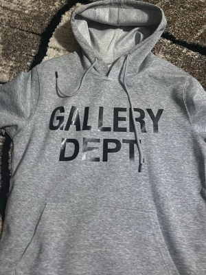 Grå hoodie från Gallery Dept - Snygg grå hoodie från Gallery Dept med svart tryck på bröstet. Klassisk modell med huva och dragsnören samt känguruficka framtill. Tillverkad i mjukt bomullsmaterial för skön känsla. Perfekt för en avslappnad streetwear-look.