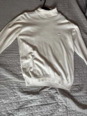 Vit stickad tröja från Lacoste - Vit stickad tröja från Lacoste med diskret krokodillogga på bröstet. Tröjan har hög krage, långa armar, väldigt clean!
