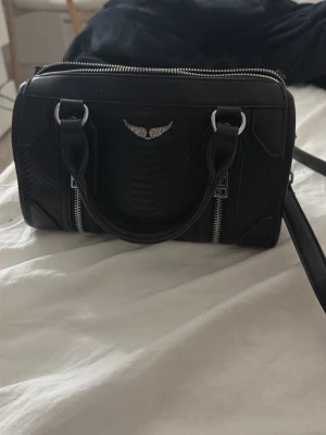 Svart handväska  - Snygg svart handväska i läderimitation med struktur och silverfärgade detaljer. Väskan har två handtag, avtagbar axelrem och två dragkedjor framtill. Dekorativt emblem med vingar på framsidan. Rymligt huvudfack med innerficka. Lite sliten på vissa ställen men syns inte mycket. Köpte för 5000 ungefär på Vinted men var inte riktigt min stil