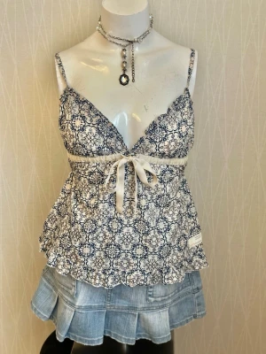 Blåvit mönstrad linnetopp med rosett - Supersöt linnetopp med tunna axelband och blått, detaljerat mönster på vit botten. Toppen har en v-ringning och en dekorativ rosett framtill under bysten. Perfekt för varma dagar och ger en romantisk vibe.