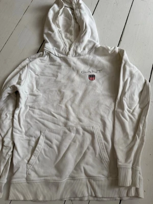 Vit hoodie från GANT med logga - Säljer en stilren vit hoodie från GANT med broderad logga och sköld på bröstet. Hoodien har en klassisk känguruficka, ribbade muddar och huva. Perfekt för dig som gillar enkel och tidlös design. Materialet känns mjukt och bekvämt.