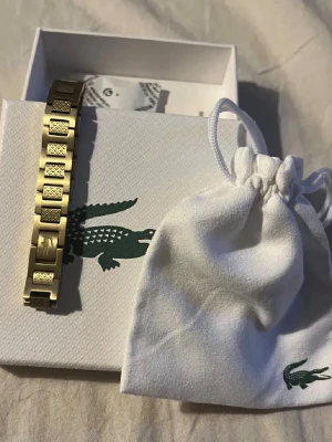 Guldigt armband från Lacoste - Snyggt guldigt armband från Lacoste med rektangulära länkar och texturerade detaljer. Armbandet har den klassiska Lacoste-loggan ingraverad på en av länkarna. Kommer i original ask och tygpåse med krokodil-logga.