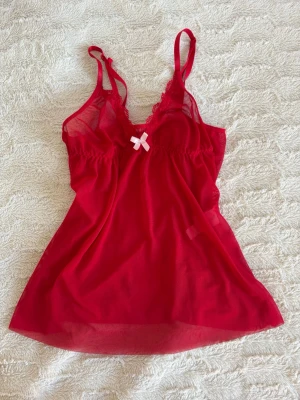 Rött nattlinne med spets  - Snyggt rött nattlinne i mesh med tunna axelband och spetsdetaljer vid bysten. Framtill sitter en liten söt vit rosett. Materialet är lätt, aldrig använd.