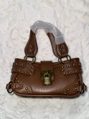 Brun handväska med nitar och låsdetalj - Cool brun handväska i skinnimitation med grov struktur, dekorerad med mässingsfärgade nitar och ett stort lås framtill. Väskan har dubbla handtag och detaljerade remmar för en edgy look. Perfekt accessoar för att lyfta din outfit.