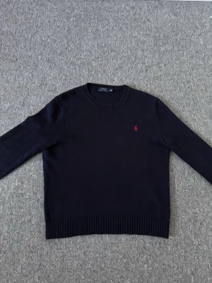 Mörkblå stickad tröja från Polo Ralph Lauren - Säljer en mörkblå stickad tröja från Polo Ralph Lauren med den klassiska röda loggan broderad på bröstet. Tröjan har rund halsringning, långa ärmar och ribbade muddar vid ärmslut och nederkant. Perfekt till jeans eller chinos.