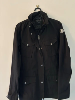 Svart vindjacka från Peak Performance - Snygg svart vindjacka från Peak Performance med silverfärgade knappar och dragkedja. Jackan har fyra fickor framtill och en patch med fjäderdetalj på ärmen. Rak passform och hög krage ger en clean look. Perfekt för blåsiga dagar. 