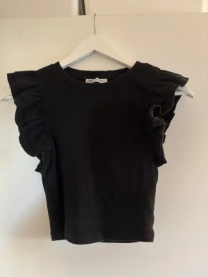 Svart volangtopp från Zara - Svart topp från Zara med volangdetaljer på axlarna och ribbad struktur. Modellen är ärmlös med rund halsringning och har en croppad passform som ger en trendig look. Perfekt att styla med jeans eller kjol.