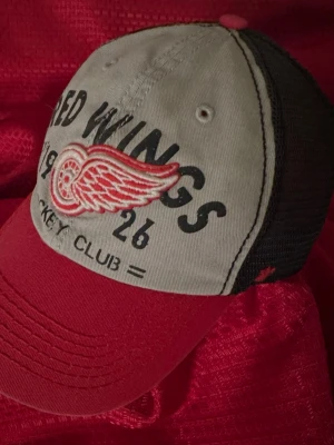 Detroit Red Wings keps röd/grå - Snygg keps med broderad Detroit Red Wings-logga framtill. Framsidan är grå med svart text och röd brodyr, skärmen är röd och baksidan är i svart mesh för extra ventilation. Justerbar passform och klassisk trucker-stil. Perfekt för hockeyfans!