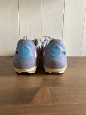 Nike TIEMPO vita fotbollsskor med blå detaljer - Säljer ett par vita Nike fotbollsskor med blå och rosa detaljer. Skorna har snörning, låg profil och tålig syntetisk ovandel. Sulan är anpassad för spel på gräs och har dobbar för bra grepp. Perfekta för dig som vill sticka ut på planen!