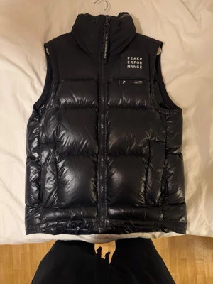 Svart dunväst från Peak Performance - Snygg svart dunväst från Peak Performance med hög krage och dragkedja framtill. Västen har en glansig finish, diskret logga på bröstet och quiltad design. Perfekt för lager-på-lager och håller dig varm under kyliga dagar.