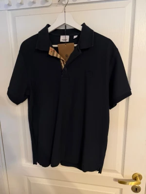 Svart pikétröja från Burberry - Klassisk svart pikétröja från Burberry med korta ärmar och den ikoniska rutiga detaljen på insidan av kragen. Tröjan har knappar framtill och diskret Burberry-logga broderad på bröstet. Perfekt för en stilren och avslappnad look.