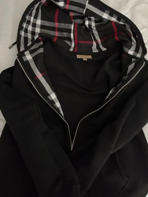 Svart hoodie från Burberry med rutig huva - Säljer en svart hoodie från Burberry med dragkedja och klassisk rutig huva i svart, vitt och rött. Hoodien har en stilren look och är tillverkad i mjukt material. Perfekt för dig som vill ha en exklusiv och trendig hoodie med ikonisk Burberry-touch. Den är gjord av ull väldigt mjuk kan sänka pris för möte i Stockholm 