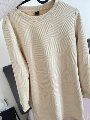 Beige mönstrad klänning med struktur - Säljer en beige klänning i medium med coolt strukturerat geometriskt mönster. Klänningen har lång ärm och rak passform, perfekt för dig som gillar stilrena men ändå unika plagg. Materialet känns mjukt och bekvämt mot huden.