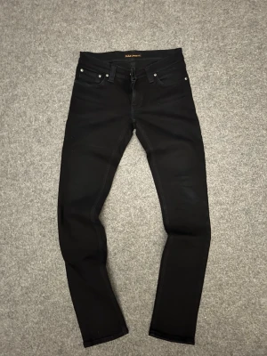 Svarta slim jeans från Nudie Jeans - Säljer ett par svarta slim jeans från Nudie Jeans med klassisk femficksmodell och smal passform. Jeansen har svarta sömmar och diskreta detaljer, tillverkade i stretchigt denim för extra komfort. Perfekta för en clean och stilren look.