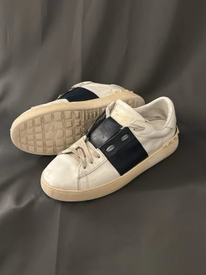 Valentino Open sneakers vit/blå skinn - Säljer ett par Valentino Open sneakers i vitt skinn med bred mörkblå rand över mitten och gulddetaljer på plösen. Klassisk låg modell med snörning och platt sula. Perfekta för dig som gillar exklusiva och stilrena sneakers.