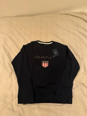 Mörkblå sweatshirt från GANT helt oanvänd  - !HELT OANVÄND!    Svart sweatshirt från GANT med broderad logga och sköld med stjärnor och ränder på bröstet. Rund halsringning med vit detalj och långa ärmar. Perfekt för en stilren och sportig look.