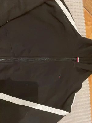 Svart hoodie från Tommy Hilfiger - Snygg svart hoodie från Tommy Hilfiger med dragkedja framtill och vita band längs ärmarna där det står 'Tommy Hilfiger Est. 1985'. Liten logga på bröstet och klassiska Hilfiger-detaljer i rött, vitt och blått. Perfekt för en stilren och sportig look.