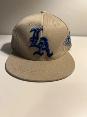 Beige LA keps med blå brodyr - Snygg beige keps med blå LA-logga broderad framtill och ett blått Los Angeles California-m tip på sidan. Klassisk rak skärm och justerbar baksida. Perfekt för dig bon gillar streetwear och vill ha en stilren accessoar.