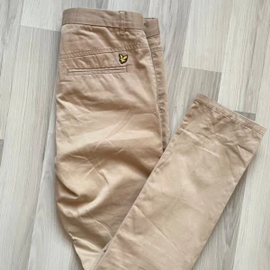 Beige chinos från Lyle & Scott - Snygga beige chinos från Lyle & Scott med klassisk passform och broderad gul logga ovanför bakfickan. Byxorna har bälteshällor, raka ben och är tillverkade i mjukt bomullstyg som ger en clean och stilren look.