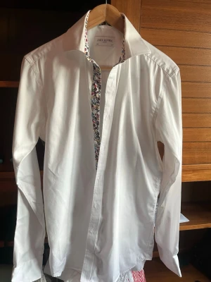 Vit skjorta John Henric - Vit skjorta från John Henric med detalj på insidan av kragen och knappslån i form av ett blommigt mönster. Skjortan har klassisk krage. Perfekt för dig som vill ha en stilren look med en twist. Nypris 1300 kr 