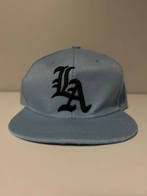 Ljusblå LA snapback keps - Snygg ljusblå snapback keps med stor svart LA-logga broderad framtill. Justerbar plastrem bak för perfekt passform. Klassisk platt skärm och ventilerande hål upptill. Perfekt accessoar för en streetwear-look.