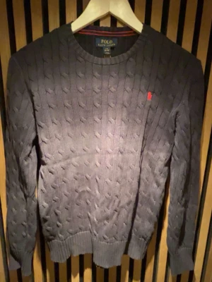 Mörkblå kabelstickad tröja Polo - Mörkblå kabelstickad tröja från Polo Ralph Lauren med rund hals och röd broderad logotyp på bröstet. Tröjan har ribbade muddar vid ärmslut och nederkant, perfekt för att hålla värmen under kyliga dagar.