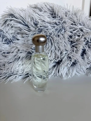 Estee Lauder Pleasures Perfume - Stilren parfymflaska i genomskinligt glas med en rund, matt silverfärgad kork. Flaskan har en mjukt böjd form och innehåller en klar vätska. Perfekt som en snygg detalj på sminkbordet eller i badrummet.