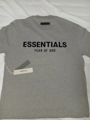 Grå Essentials Fear of God t-shirt - Snygg grå t-shirt från Essentials Fear of God med svart tryck framtill. Klassisk passform och rund halsringning, perfekt för en stilren och avslappnad look. Tillverkad i mjuk bomull för maximal komfort.