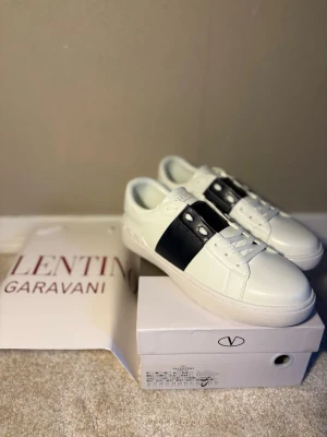 Valentino Garavani vita sneakers - Snygga vita sneakers från Valentino Garavani med mörkblå läderdetalj och nitar på sidan. Klassisk låg modell med rund tå och vit sula. Perfekta för dig som gillar stilrena och exklusiva skor med en edgy touch.