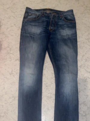 Blå jeans från Nudie Jeans - Säljer ett par klassiska blå jeans från Nudie Jeans med snygg slitning och kontrastsömmar. Jeansen har fem fickor, normal midja och raka ben. Tillverkade i denim med en cool tvätt och Nudie-logga på bakfickan.
