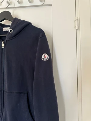 Mörkblå hoodie från Moncler - Snygg mörkblå hoodie från Moncler med dragkedja framtill och fickor. Huva med vita och röda detaljer samt Moncler-logga på ärmen. Tillverkad i mjukt bomullsmaterial, perfekt för en avslappnad och trendig look.