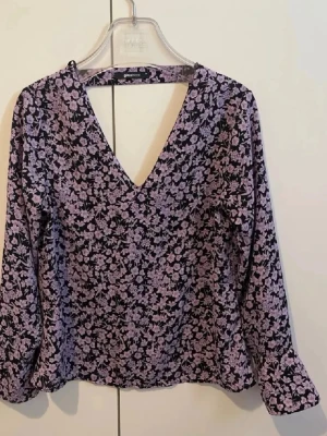 Lila blommig blus från Gina Tricot - Snygg blus från Gina Tricot i svart med lila blommönster. Blusen har djup V-ringning och långa ärmar. Materialet känns mjukt och lätt, perfekt för en trendig look. Passar till både jeans och kjol för en cool stil.