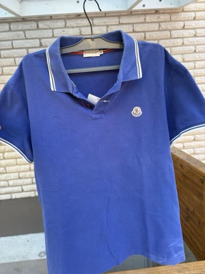 Blå pikétröja från Moncler - Snygg blå pikétröja från Moncler med vitrandiga detaljer på krage och ärmslut. Tröjan har klassisk pikékrage, korta ärmar och Moncler-logga på bröstet. Tillverkad i bomull för en bekväm och luftig känsla. Storlek XL men är som en L eftersom Moncler storlekarna är små. 
