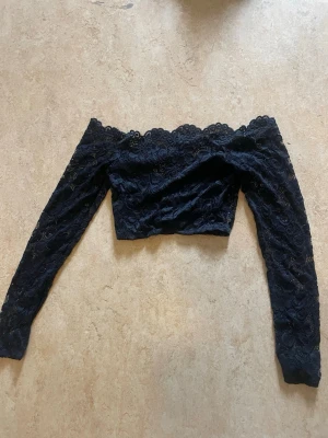 Svart off shoulder spets-crop top - Trendig svart crop top i spets med off shoulder-modell och långa ärmar. Toppen har en romantisk känsla med vacker spetsdetalj och figurnära passform. Perfekt för att skapa en snygg och ungdomlig look.