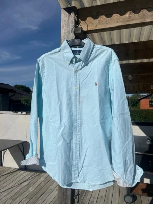 Ljusblå skjorta med knappar - Snygg ljusblå skjorta med klassisk krage och knappar framtill. Skjortan har långa ärmar och en liten broderad logga på bröstet. Perfekt för en stilren look och passar till många olika outfits.