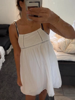 Vit sommarklänning med spetsdetaljer - Söt vit kortklänning med tunna axelband och knyt på axlarna. Klänningen har broderade blommor och spetsdetaljer framtill, samt en luftig passform. Perfekt för varma dagar och ger en romantisk vibe.