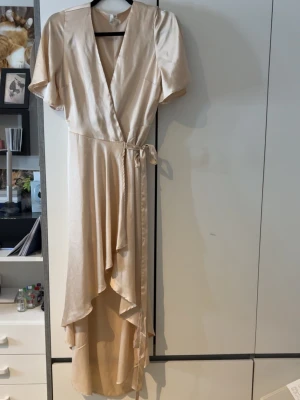 Champagnefärgad omlottklänning NLY Trend - Säljer en elegant champagnefärgad långklänning från NLY Trend. Klänningen har omlottmodell med v-ringning, korta ärmar och ett knytband i midjan. Materialet har en sidenliknande finish som ger ett lyxigt intryck. Perfekt för dig som vill sticka ut!