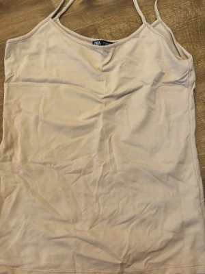 Beige linnetopp med smala axelband - Säljer en enkel och stilren beige linnetopp med smala axelband. Toppen har en rak passform och är perfekt att matcha med jeans eller kjol. Materialet känns mjukt och lätt, vilket gör den skön att bära under varma dagar.