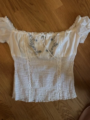 Vit offshoulder topp från Kimchi Blue - Supersöt vit offshoulder topp från Kimchi Blue med broderade blommor och spetsdetaljer. Toppen har smockad midja, puffärmar och knyt framtill. Perfekt för en romantisk look och passar till både jeans och kjol.