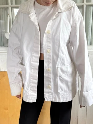 Vit oversized jeansjacka - Snygg vit jeansjacka med oversized passform och stora framfickor. Jackan har klassisk krage, knäppning med fyra knappar och breda ärmslut med knappdetalj. Perfekt för en clean och stilren look.
