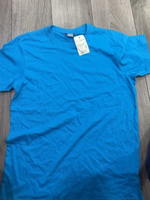 Blå basic t-shirt i bomull - En enkel och stilren blå t-shirt i mjuk bomull. T-shirten har rund halsringning och korta ärmar, perfekt för en avslappnad look. Passformen är normal och tyget är lätt och skönt mot huden.