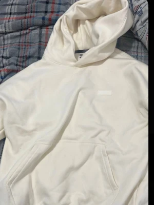 Vit hoodie med huva och magficka - Säljer en stilren vit hoodie med stor huva och klassisk magficka framtill. Hoodien har en enkel design utan synliga tryck eller mönster, vilket gör den lätt att matcha med olika outfits. Perfekt för dig som gillar en clean och avslappnad look.