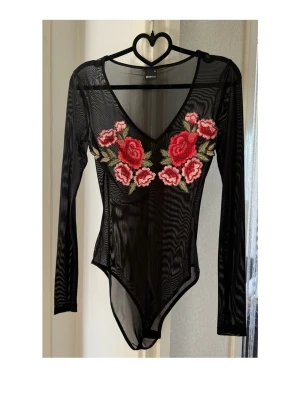 Svart meshbody med rosbroderi - Snygg svart meshbody med långa ärmar och djup V-ringning. Framsidan pryds av stora broderade rosor i rött och rosa med gröna blad. Materialet är genomskinligt mesh som ger en trendig look. Perfekt för dig som vill sticka ut med detaljerade broderier.